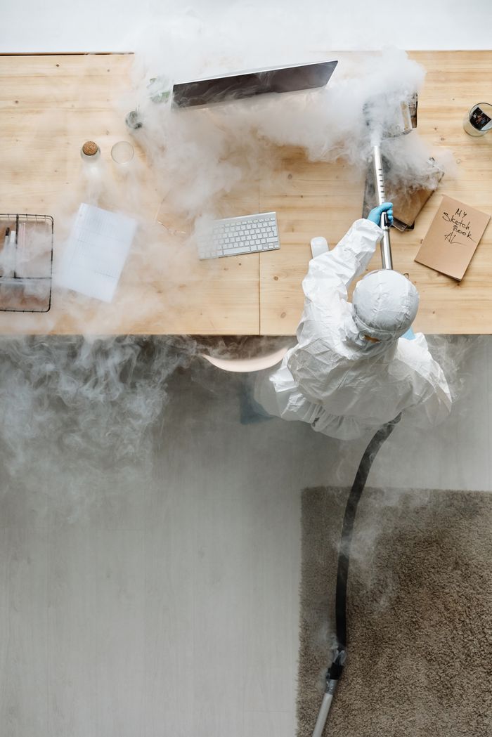 A person wearing protective gear is sanitizing an office desk with fumigation equipment in daylight.